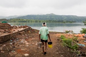 Nasra Solanki, 50, walks past demolished homes to fill water from the river at Kakrana, Alirajpur, Madhya Pradesh. Before the Sardar Sarovar Dam gates shut on June 17, the village extended all the way till the green strip of land seen in the distance, he said. September 12, 2017.