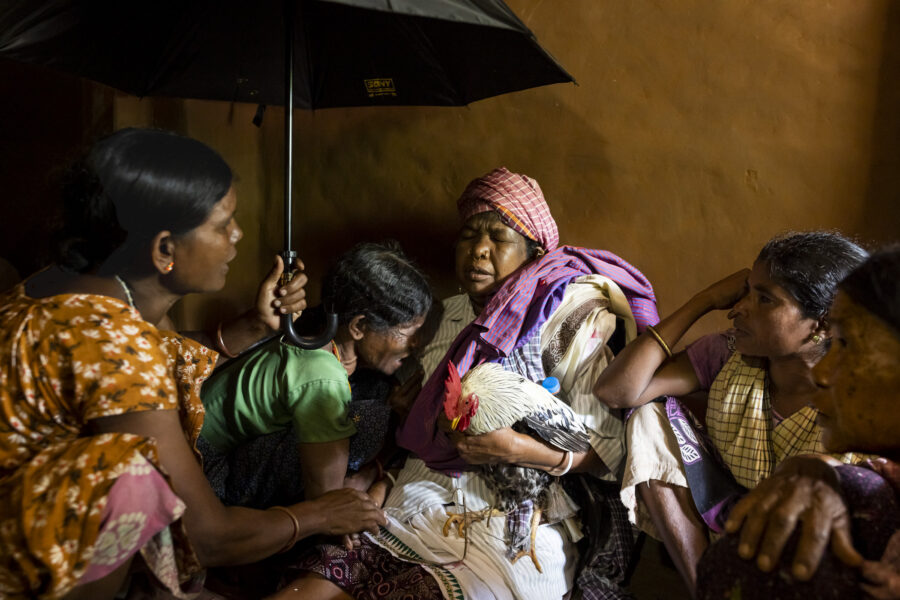 Sora shaman Lokami channels Sakana Soboro, who passed away three months ago after a brief illness and now speaks through her with his relatives. The chicken, the clothes on his shoulder and umbrella are all offerings from his family. Jorrepi, Odisha, India.